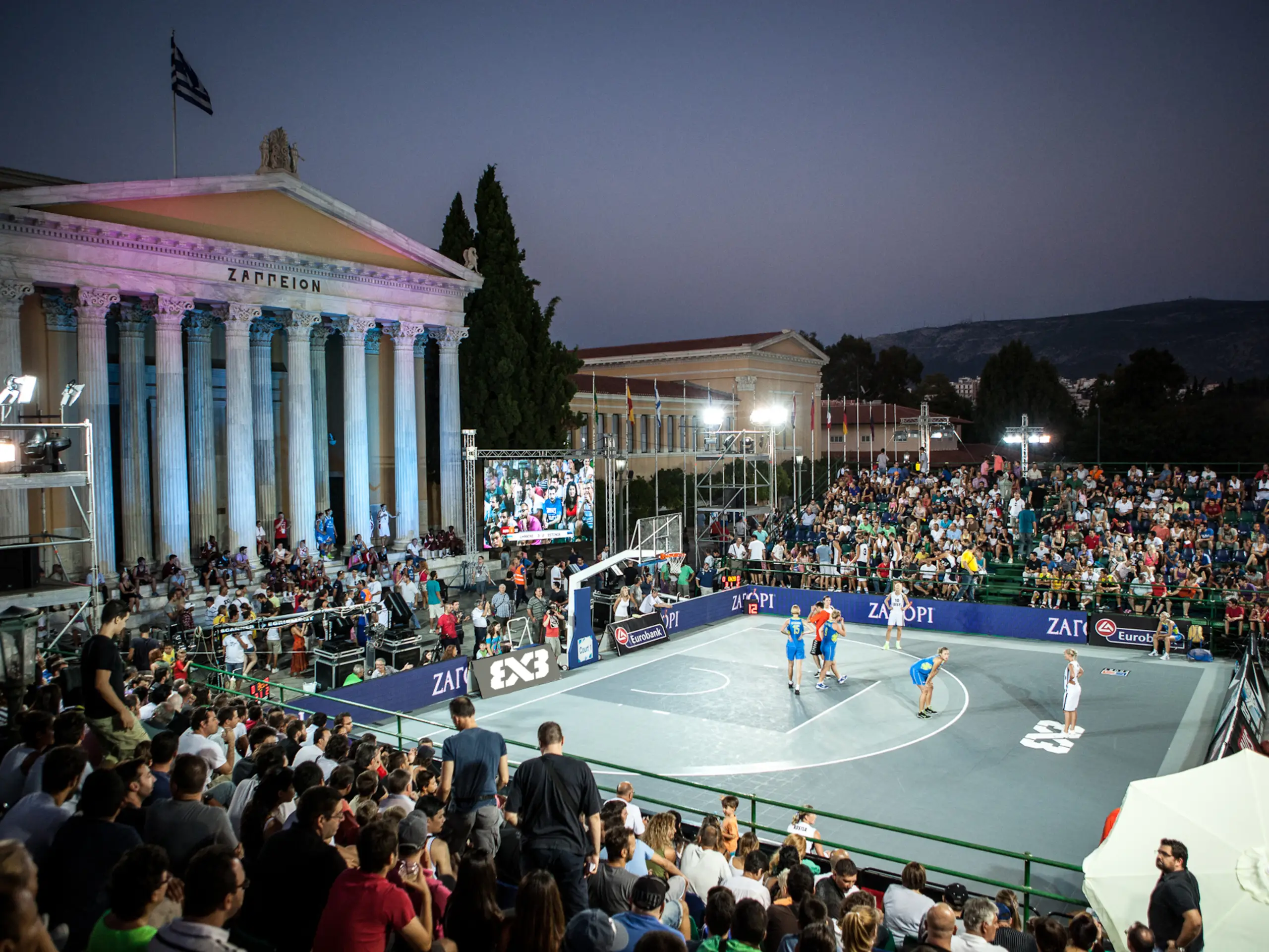 Acción de baloncesto 3x3 al aire libre con un jugador atacando al aro y público alrededor de la pista urbana
